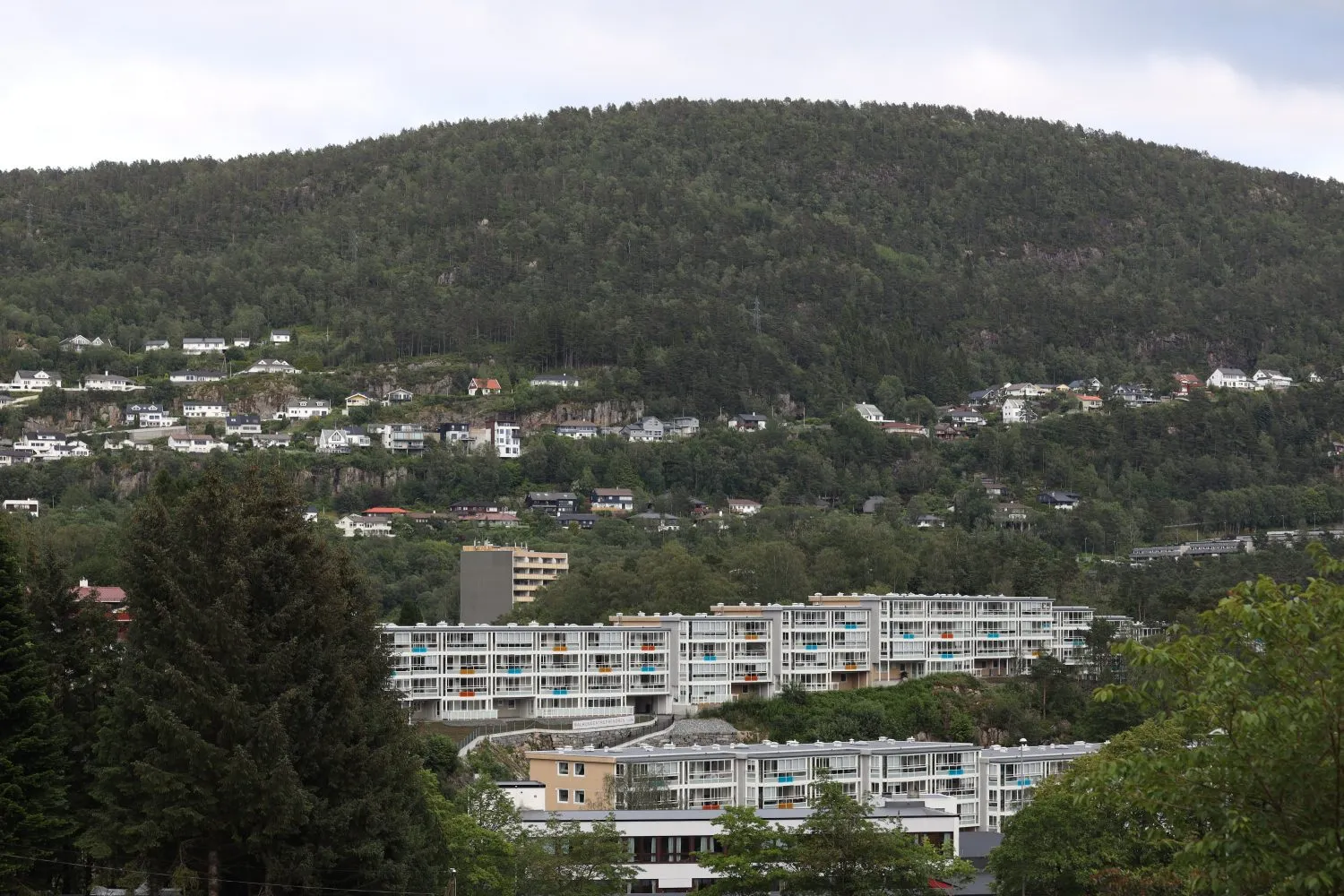 To lavblokker i fire etasjer over hverandre, med fargespill på terrassene, foran en skogkledd li i Fyllingsdalen
